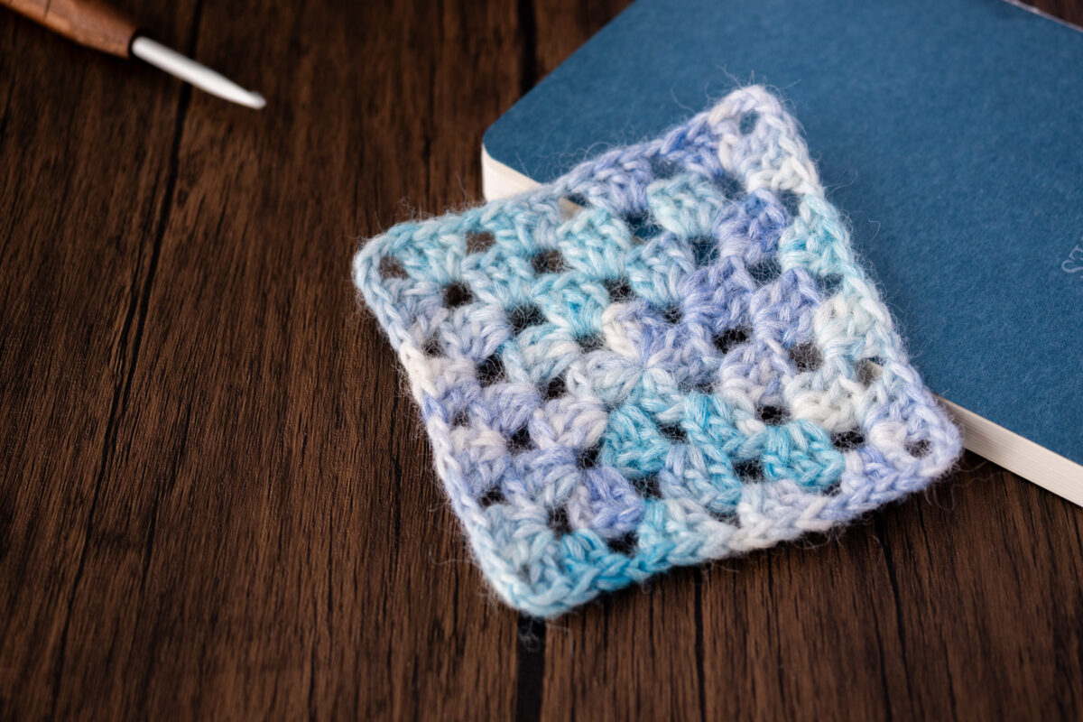 Ein blau-weißes Granny Square, gehäkelt ohne Farbwechsel, liegt auf einem geschlossenen blauen Notizbuch auf einer Holzoberfläche. Im Hintergrund ist teilweise eine Häkelnadel sichtbar.