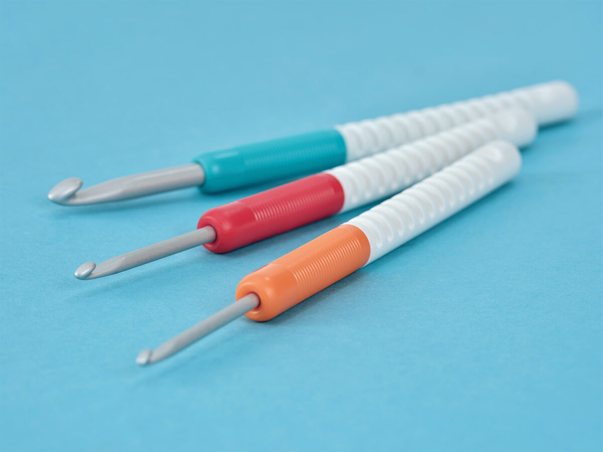 Beste Häkelnadel für Anfänger zum Häkeln lernen Auf einer hellblauen Fläche sind drei Häkelnadeln mit weißen, strukturierten Griffen und farbigen Griffen (türkis, rot und orange) angeordnet, ideal als Häkelnadel für Anfänger, wobei die Metallhakenenden nach vorne zeigen.