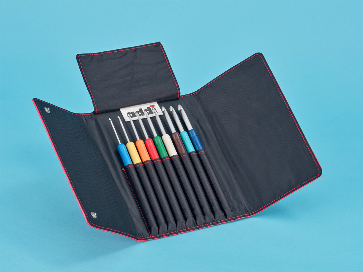 Beste Häkelnadel für Anfänger zum Häkeln lernen Ein schwarzes Etui mit roter Naht enthält acht Häkelnadeln für Anfängern, jede mit einem andersfarbigen Griff, der auf einem blauen Hintergrund dargestellt ist. Der geöffnete Koffer zeigt alle Haken ordentlich organisiert im Inneren.