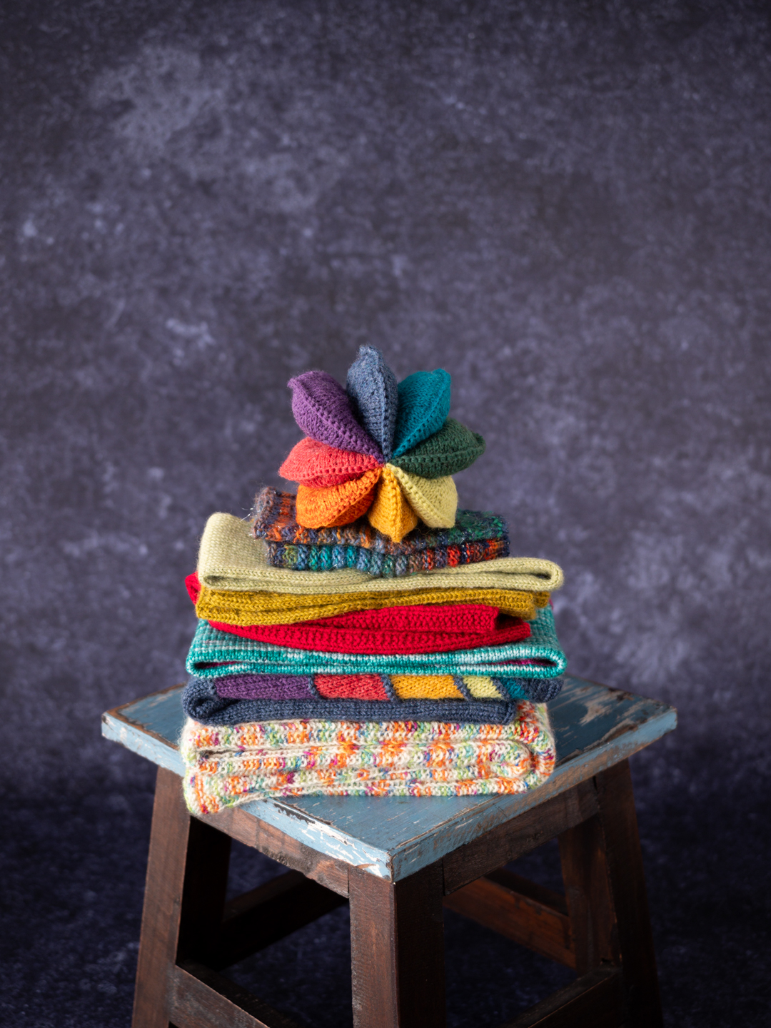 Stricken mit Sockenwolle für die Kleinen Ein Stapel von bunten, aus Sockenwolle gestrickten Kindersachen liegt auf einem Hocker.
