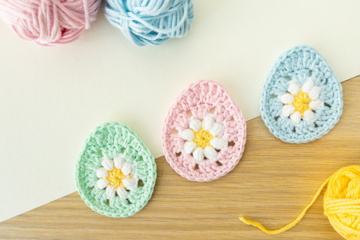 Drei gehäkelte Ei-Formen - perfekt als Osterei-Applikation häkeln - in Pastellgrün, Rosa und Blau mit Blumen in der Mitte sind neben Knäueln aus rosa, blauem und gelbem Garn auf einer hölzernen und cremefarbenen Oberfläche ausgestellt.