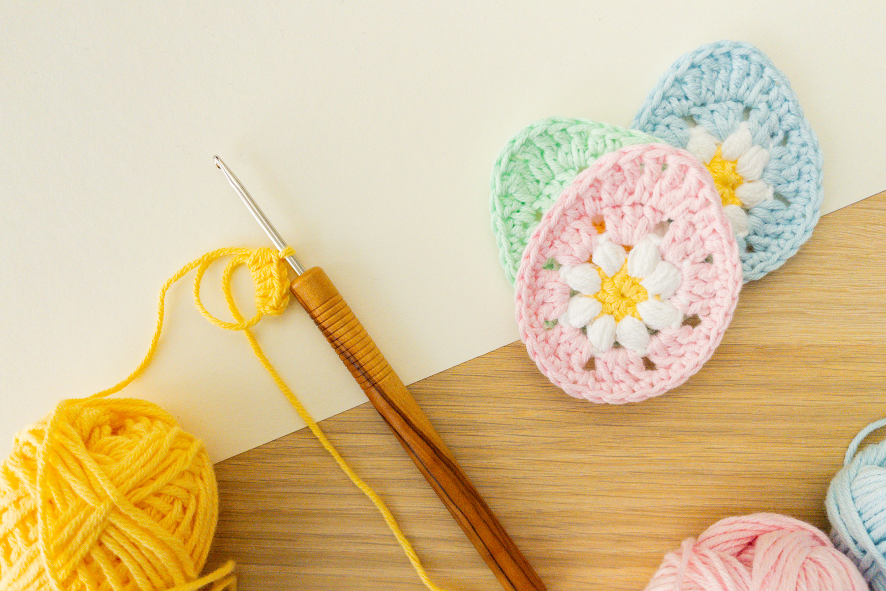 Eine Häkelnadel mit Holzgriff, gelbes Garn und farbenfrohe Häkelblumenmotive in Rosa, Blau und Grün sind auf einem hellen Holz- und cremefarbenen Hintergrund arrangiert - perfekt für Ihr nächstes osterei applikation häkeln Projekt.