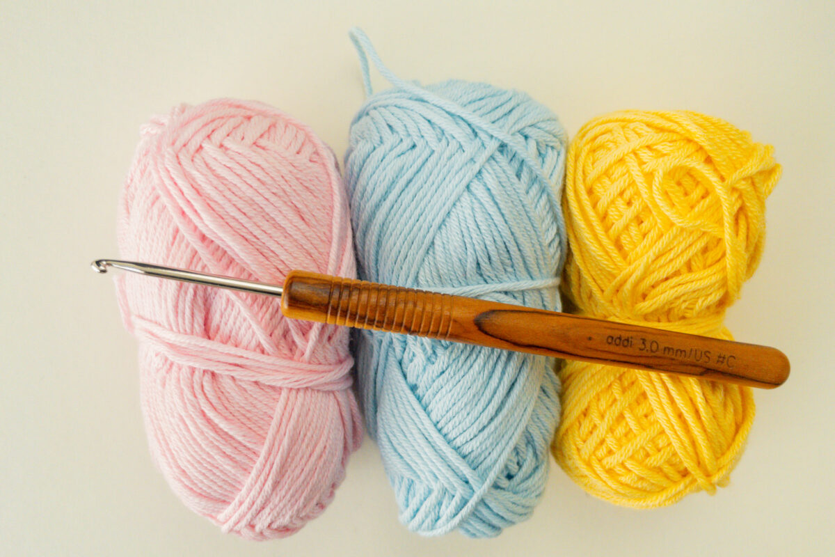 Drei Garnstränge in den Farben Rosa, Blau und Gelb liegen nebeneinander, darauf eine Häkelnadel mit Holzgriff - perfekt für Ihr nächstes Osterei-Anhänger-Häkeln oder Osterei-Applikation-Häkeln-Projekt.