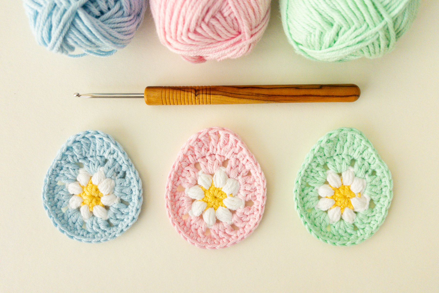 Drei gehäkelte Ostereier in Blau, Rosa und Grün mit weißen und gelben Gänseblümchenmustern sind unter einer hölzernen Häkelnadel und pastellfarbenen Garnkugeln aufgereiht - perfekt, um eine Osterei-Applikation zu häkeln oder festliche Anhänger zu gestalten.