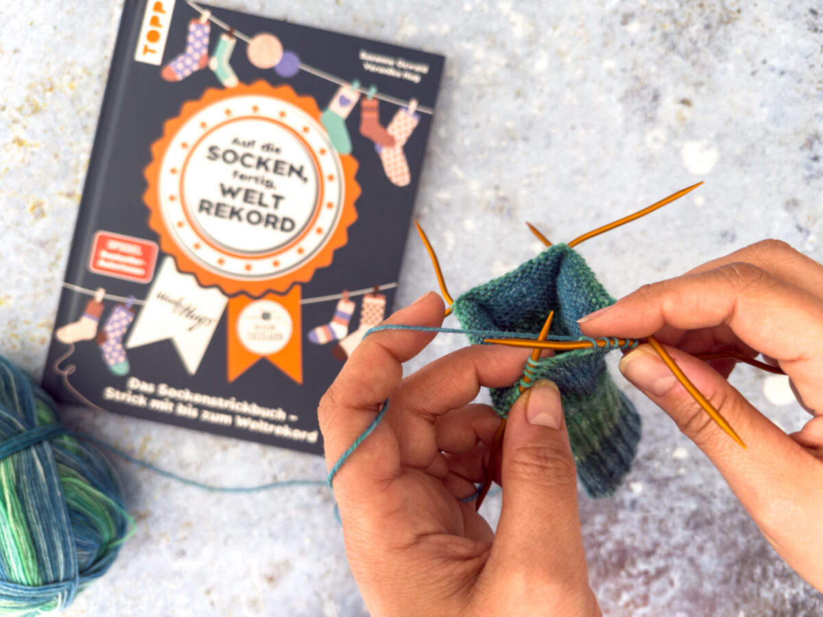 Eine Person strickt mit blau-grünem Garn und doppelspitzigen Nadeln. Im Hintergrund feiert das deutsche Strickbuch "Auf die Socken, fertig, Weltrekord" mit illustrierten Socken auf dem Cover den Sockenweltrekord.