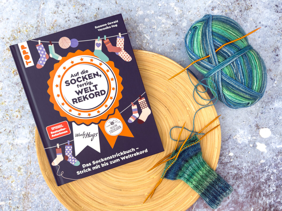 Ein deutsches Sockenstrickbuch liegt auf einer Holzplatte neben blaugrünem Garn und doppelspitzigen Nadeln mit einer teilweise gestrickten Socke, die an den Geist des Sockenweltrekords erinnert. Der Hintergrund ist eine strukturierte, gesprenkelte graue Oberfläche.
