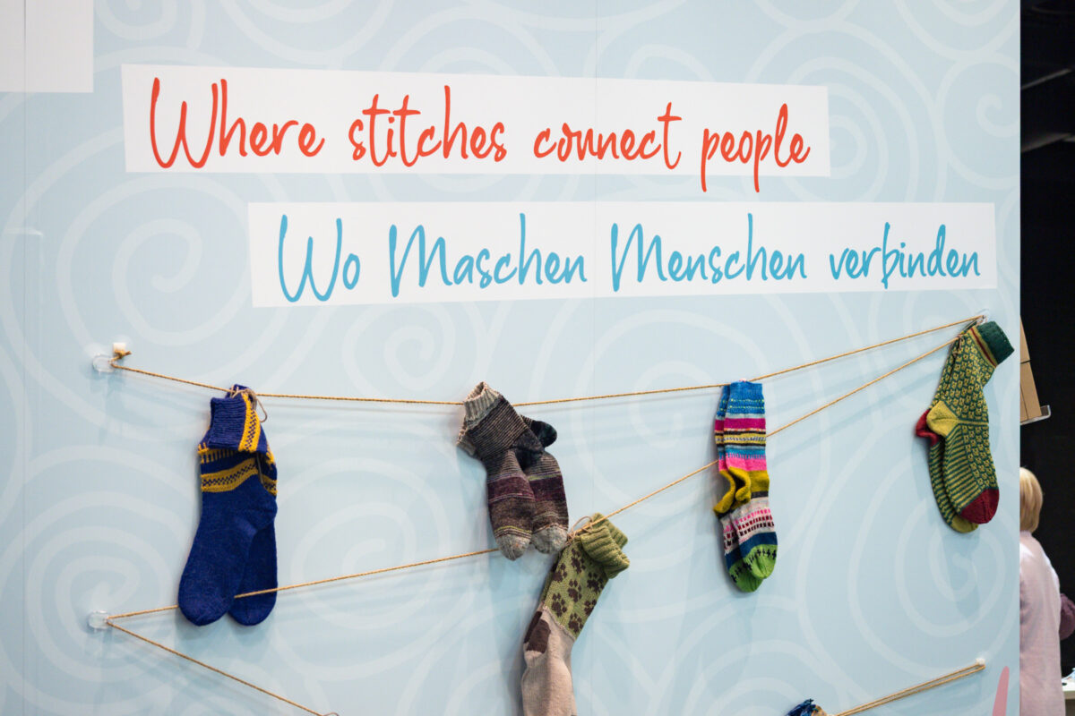 Bunte gestrickte Socken hängen an einer Schnur unter einem Schild mit der Aufschrift "Wo Maschen Menschen verbinden" und "Wo Maschen Menschen verbinden" vor einem hellblau gemusterten Hintergrund - und zelebrieren den Geist des Sockenweltrekords.