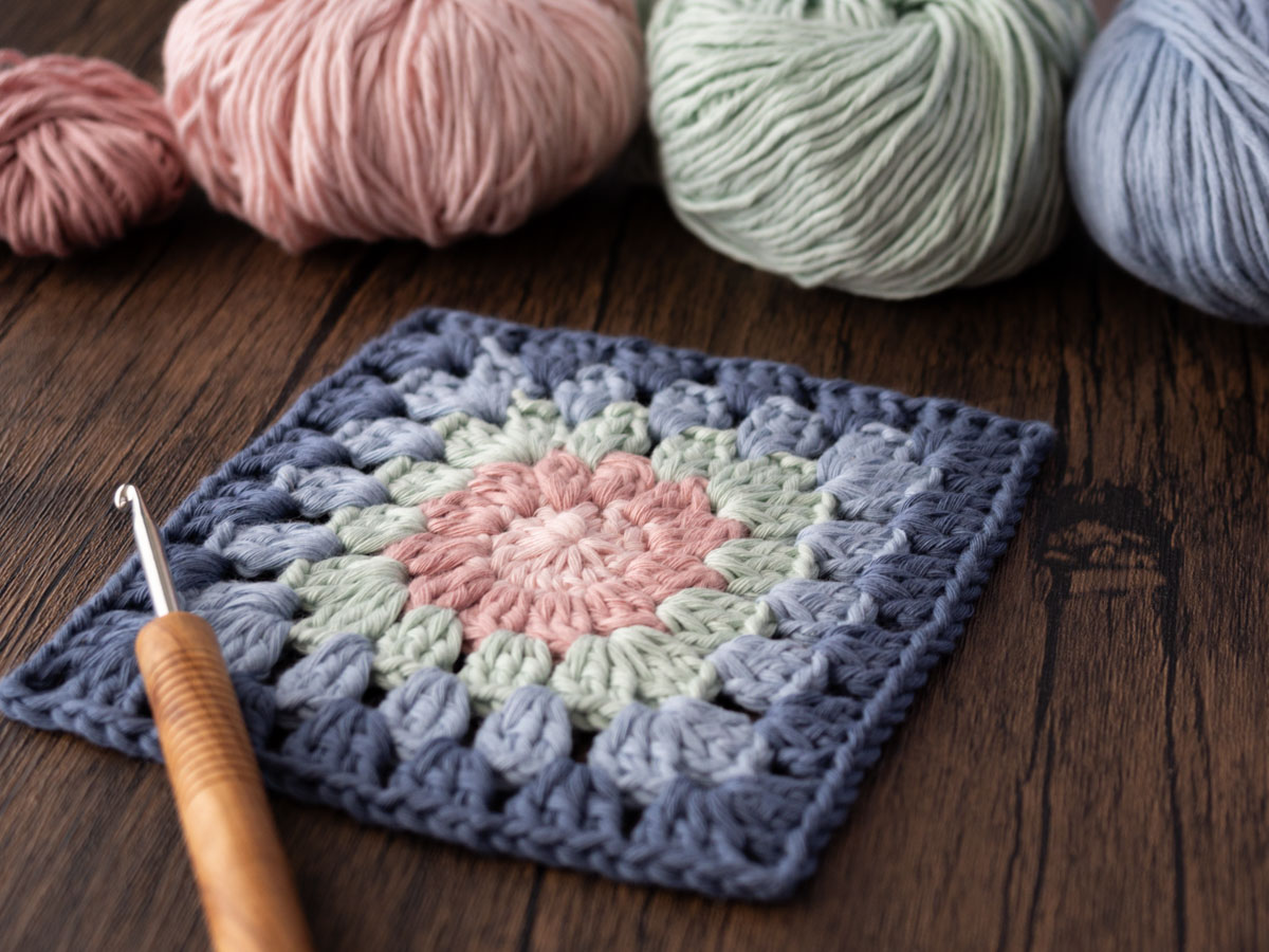 Eine Häkelnadel liegt auf einem Granny Square aus rosa, salbeigrünem und blauem Garn, daneben liegen passende Garnknäuel für alle, die nach kostenlosen Häkelanleitungen für Granny Squares suchen.